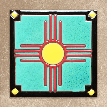 Decorative tile with a red and yellow zia symbol on a turquoise background, framed by black border with gold accents.