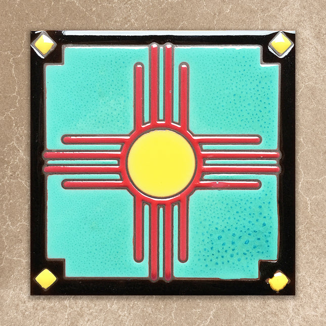 Decorative tile with a red and yellow zia symbol on a turquoise background, framed by black border with gold accents.