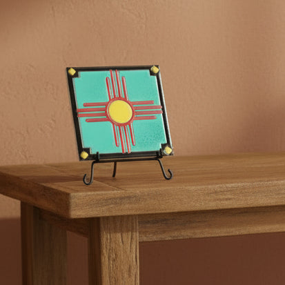 Decorative tile with a red and yellow zia symbol on a turquoise background, framed by black border with gold accents.