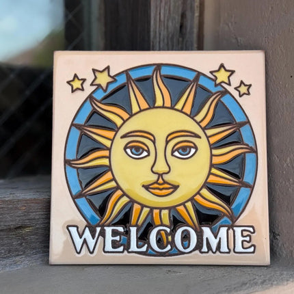 Decorative tile with sun and moon design and 'WELCOME' text on a wooden surface.